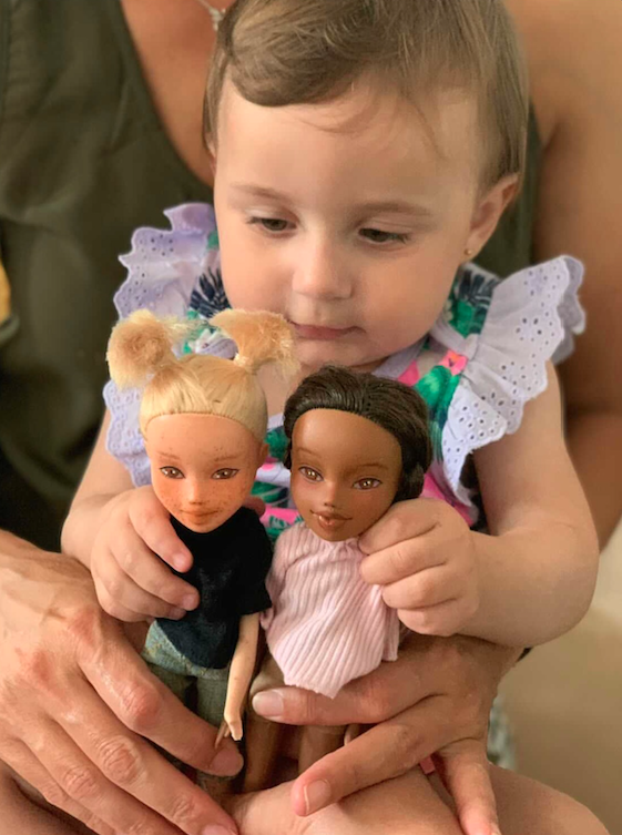 Toddler cradling two handmade dolls with different skin tones — one light, one dark
